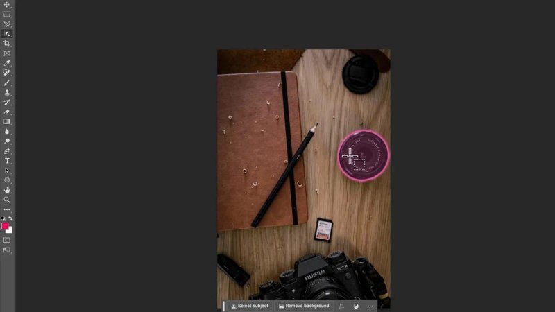 Flat lay photo on a wooden desk in Photoshop showing a notebook, pencil, camera body, and memory card, with a circular lens cap highlighted in magenta by the Object Finder crosshair cursor.