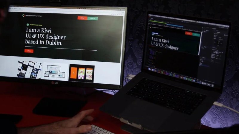 Physical dual-screen workstation showing a large external monitor rendering the full portfolio website next to an open laptop running code in Dreamweaver